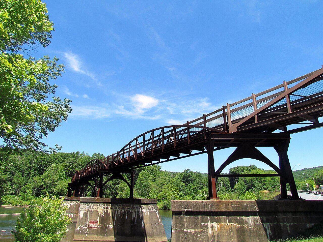 A rusty steel trestle bridge carries a trail over a river between green wooded banks