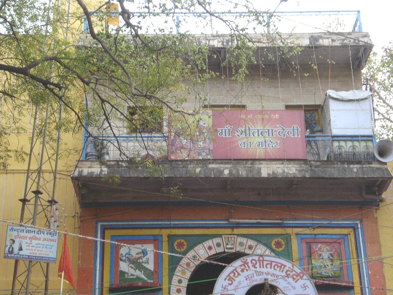 The front of a temple building with a yellow facade, a painted arched gateway and a pink sign in Hindi