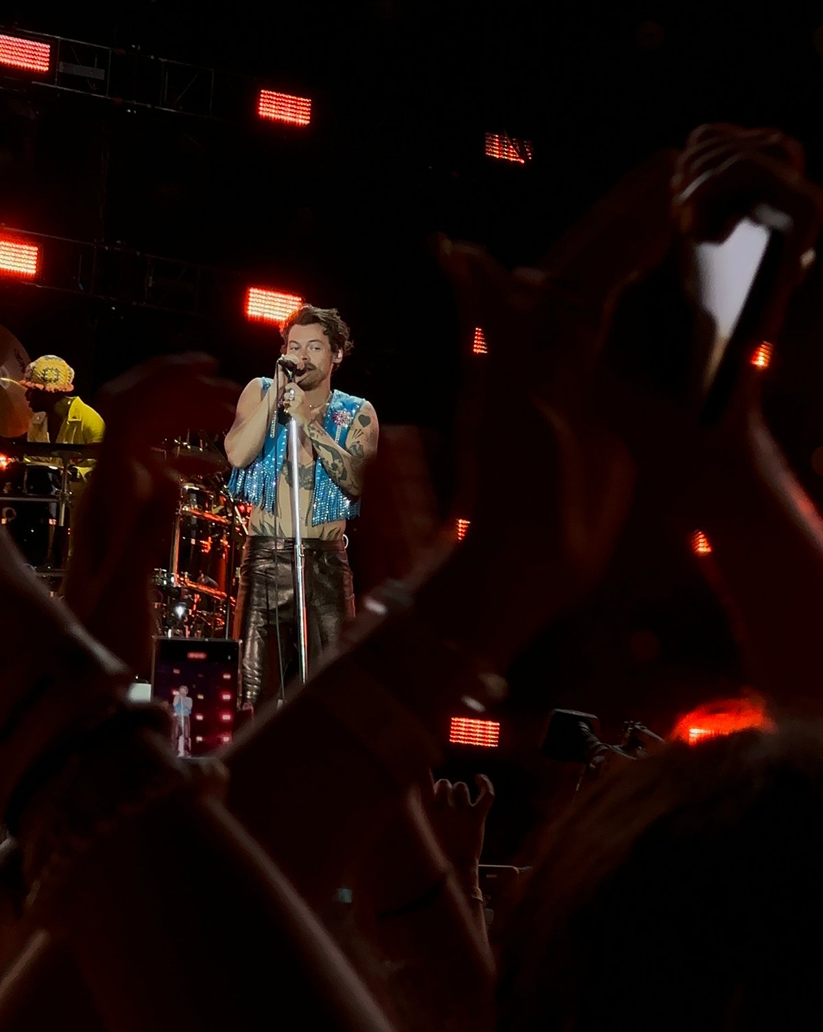 Harry Styles sings into a microphone on a dark stage lit by red lights as fans raise their hands