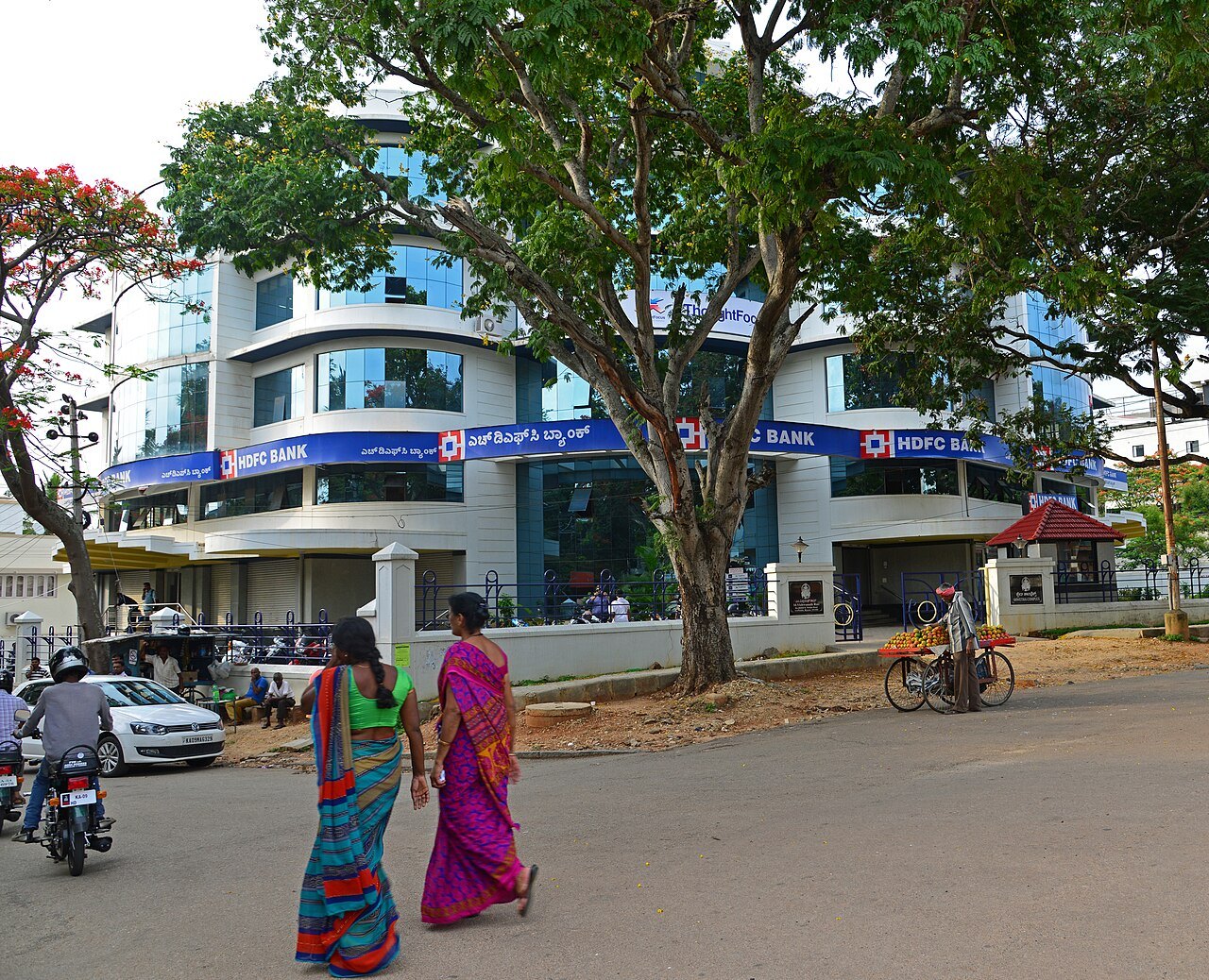 An HDFC Bank branch with a blue sign behind trees, with two women walking past