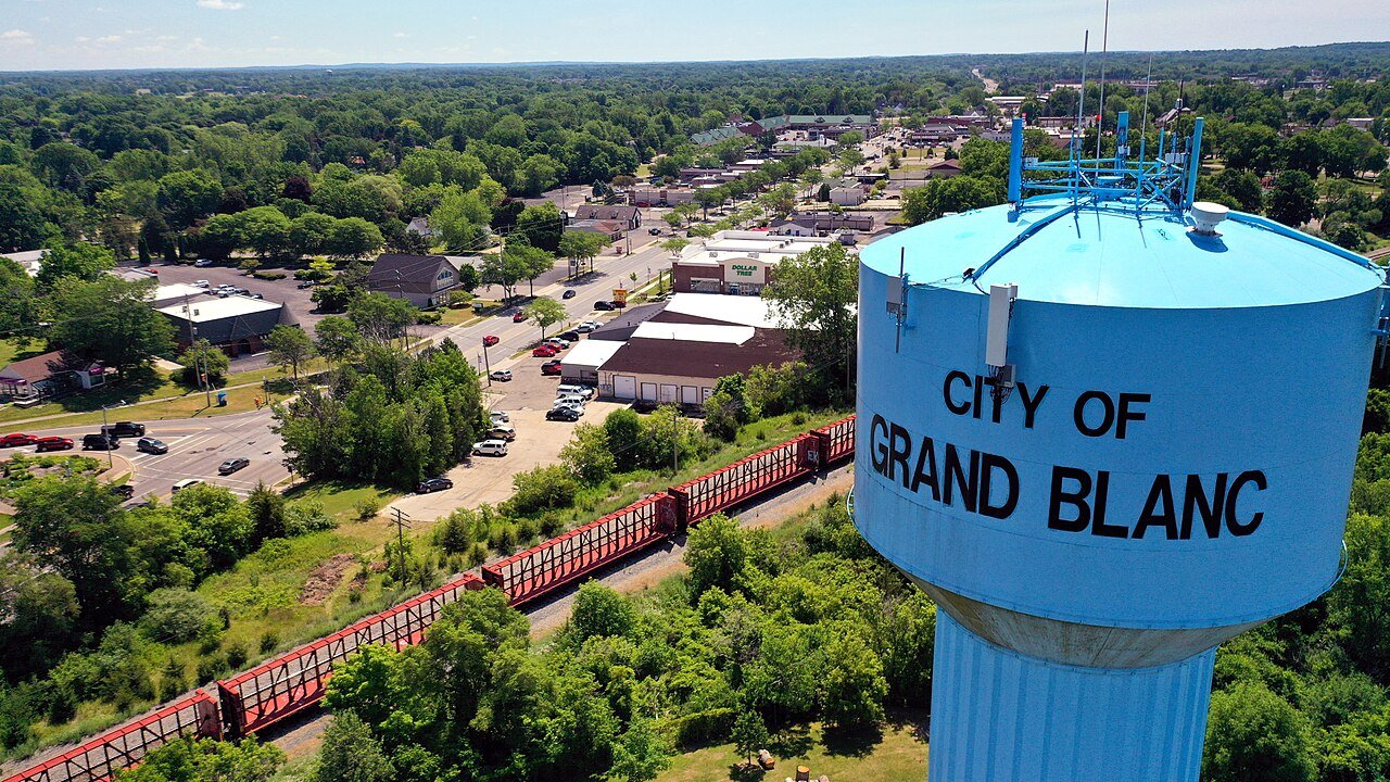 A blue water tower reading City of Grand Blanc stands above a small town with trees and a railway line