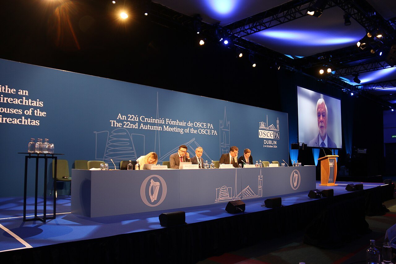 A white-haired man in a suit appears on a large video screen beside a blue-lit stage where a panel sits at a long table