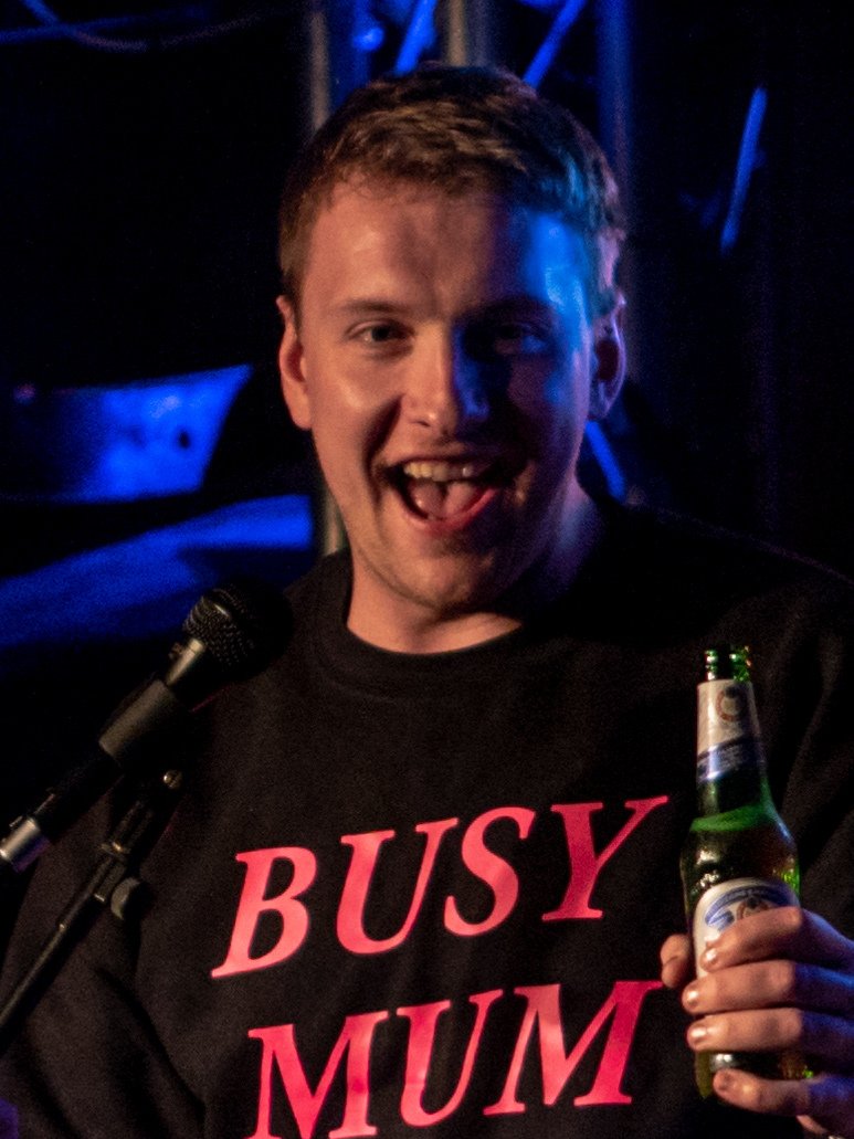 Joe Lycett, smiling with his mouth open, holds a beer on a stage lit in blue and wears a black top with pink "BUSY MUM" lettering