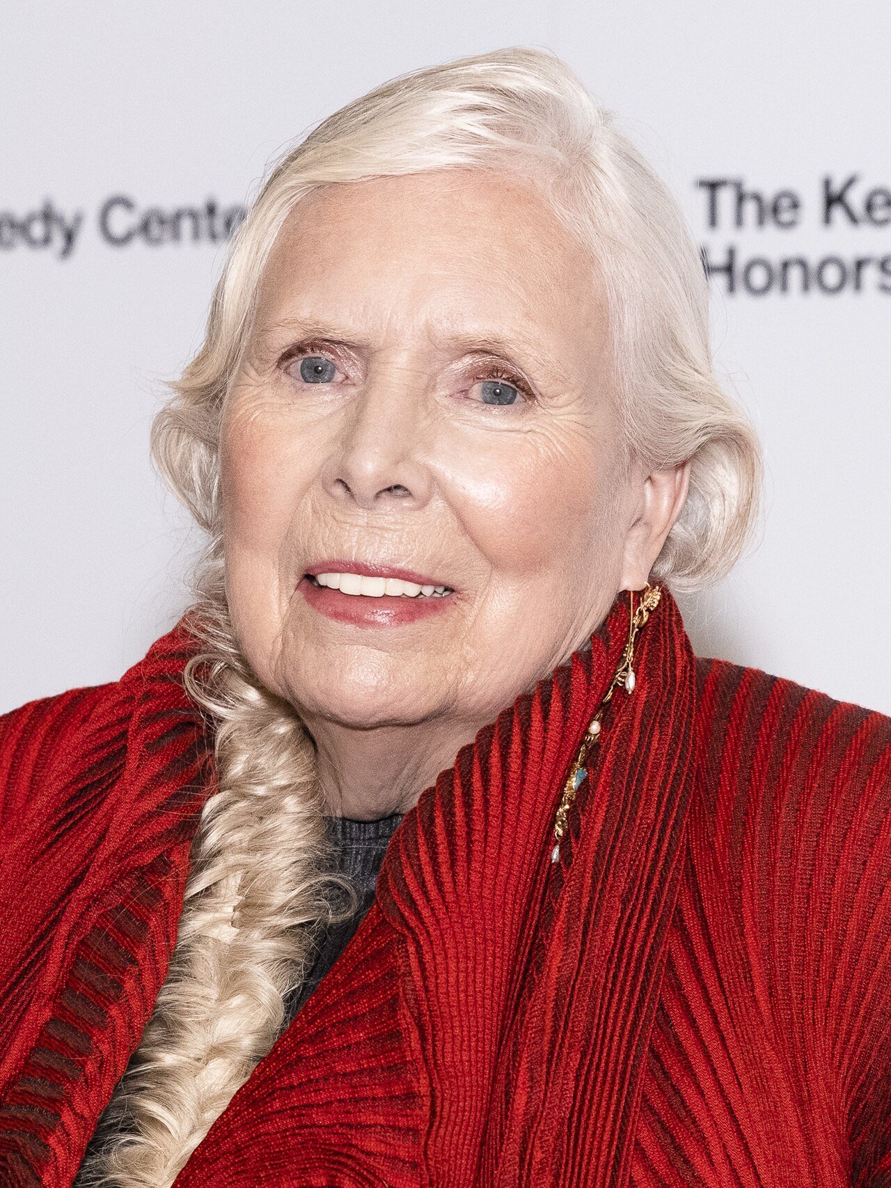 Joni Mitchell, with long white hair in a braid and a red jacket, smiles in front of a Kennedy Center Honors backdrop