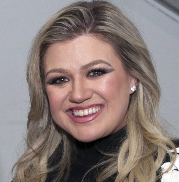 Kelly Clarkson, with long blonde hair, smiles in a black top against a grey background