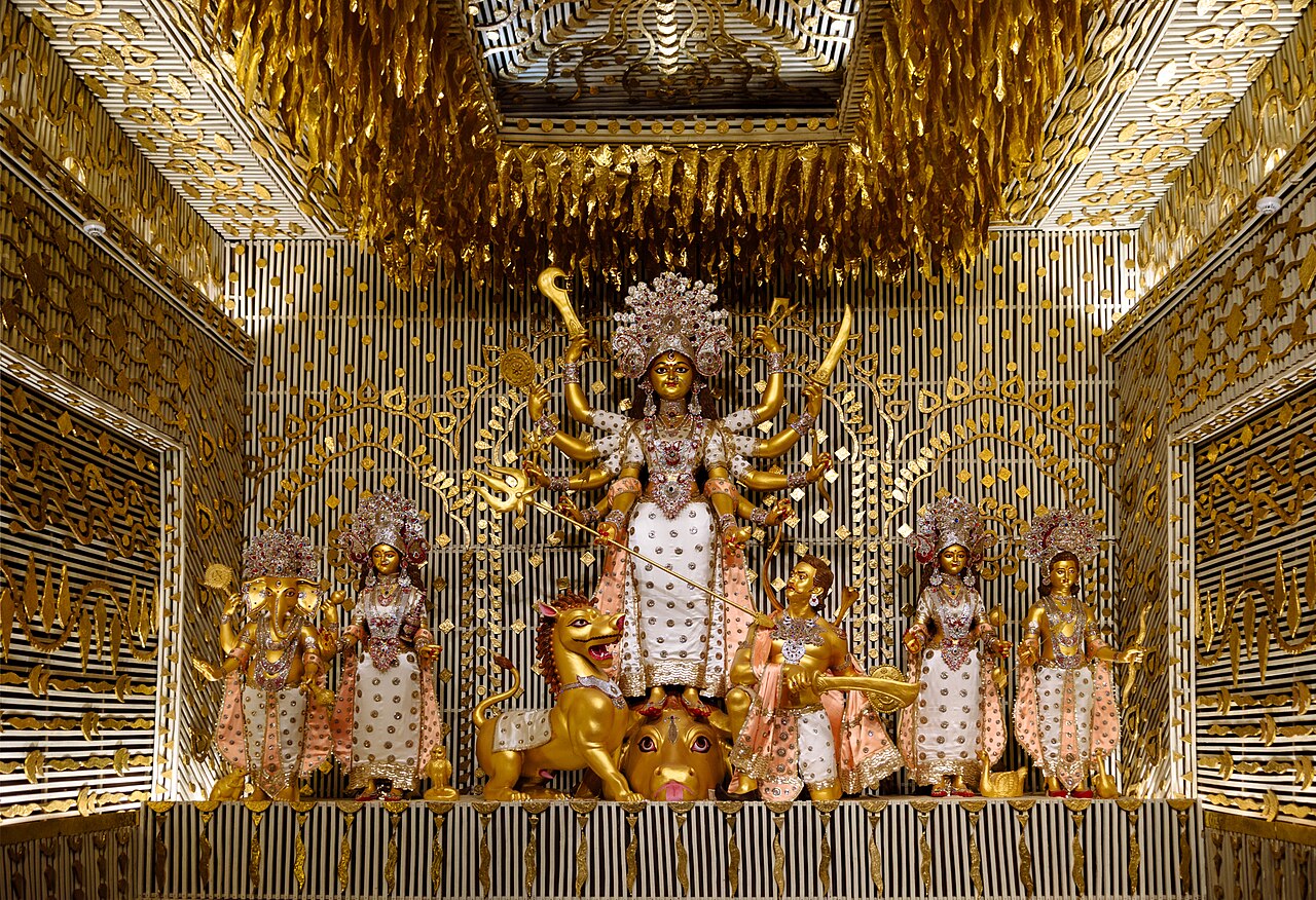 A brightly lit, gold-decorated shrine with a many-armed goddess statue at its center, flanked by other figures