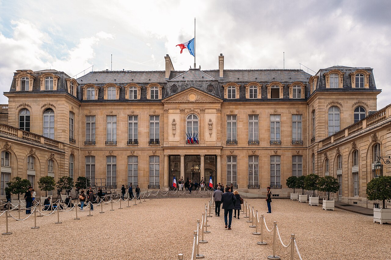 The stone front of the Élysée Palace with a French flag on the roof and people walking across a gravel courtyard lined with rope barriers