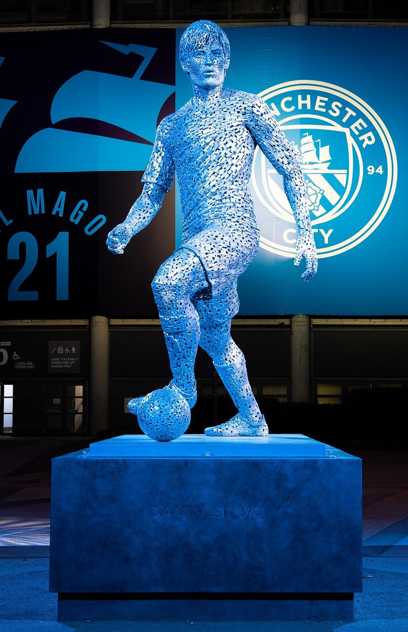 A blue statue of footballer David Silva on a plinth outside Etihad Stadium, with a large Manchester City crest behind it