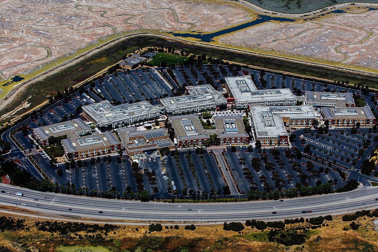 An aerial view of a large office campus of low connected buildings around a central courtyard, surrounded by parking lots and a highway beside salt marshes