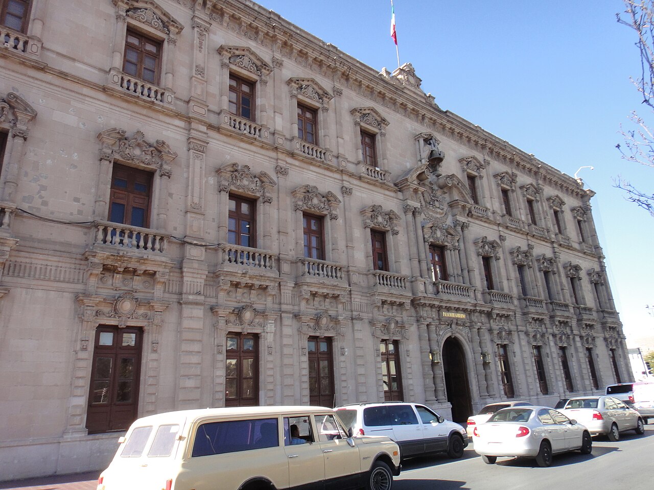 A long two-story stone government building with arched windows and a flag on the roof, with cars parked in front