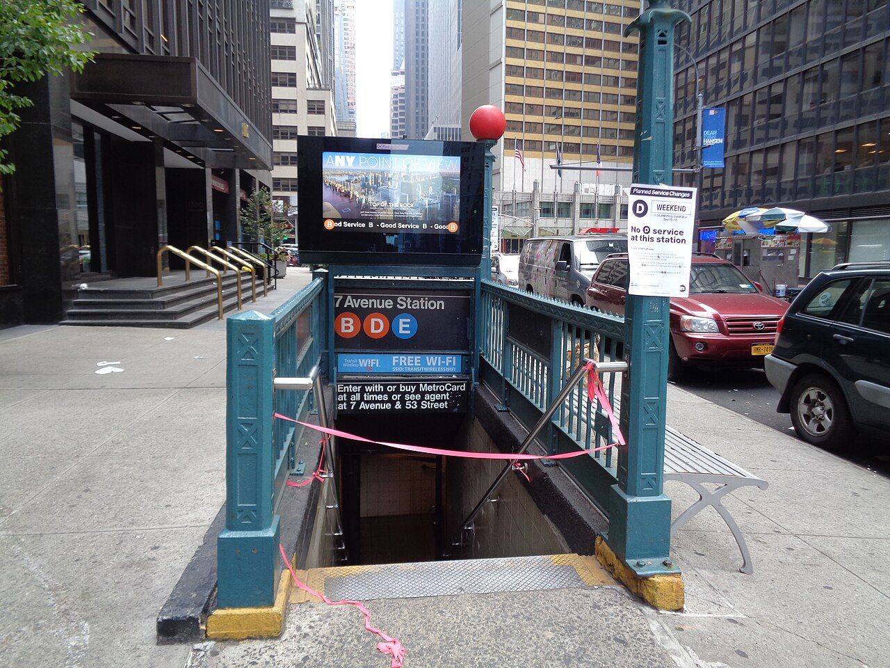 A green metal subway entrance with railings leads down a stairway on a city sidewalk, with a sign reading 7 Avenue Station and B, D and E train symbols, tall office buildings and parked cars behind it