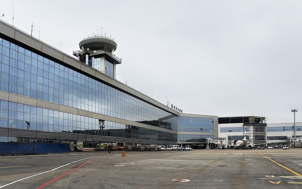 A long glass-fronted airport terminal with a round control tower on its roof stands beside an empty apron under a gray sky, with a passenger jet parked at a gate on the right