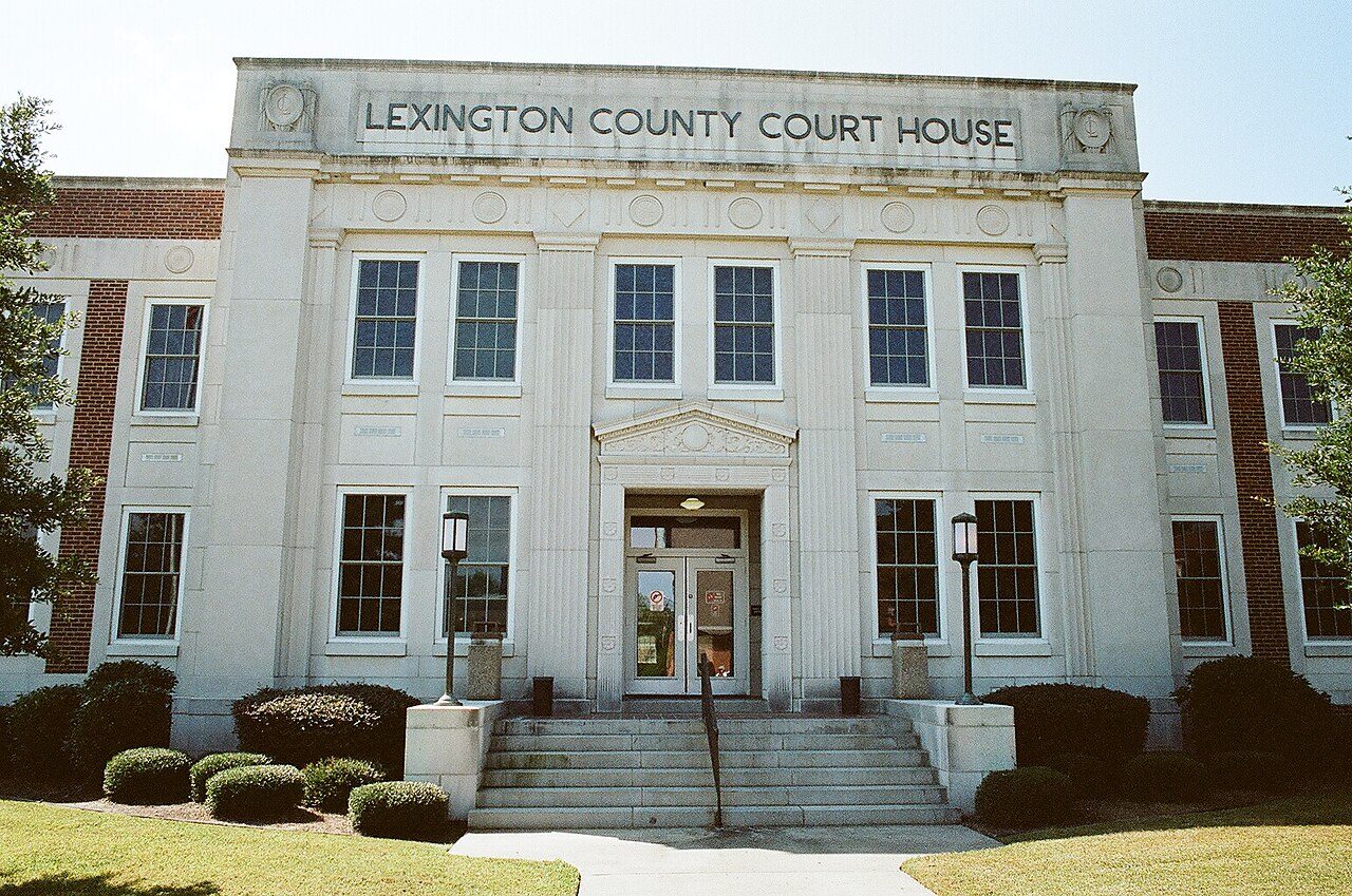 A pale stone courthouse with the words Lexington County Court House carved above tall windows and a central doorway, with steps, lamps and trimmed shrubs in front