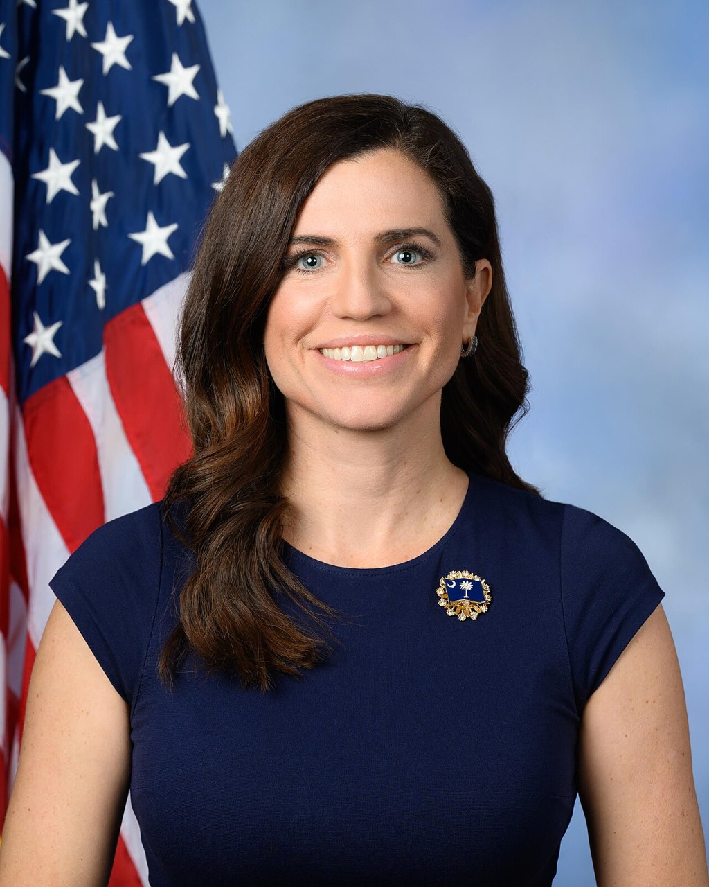 Nancy Mace in a navy dress, smiling, in front of a US flag