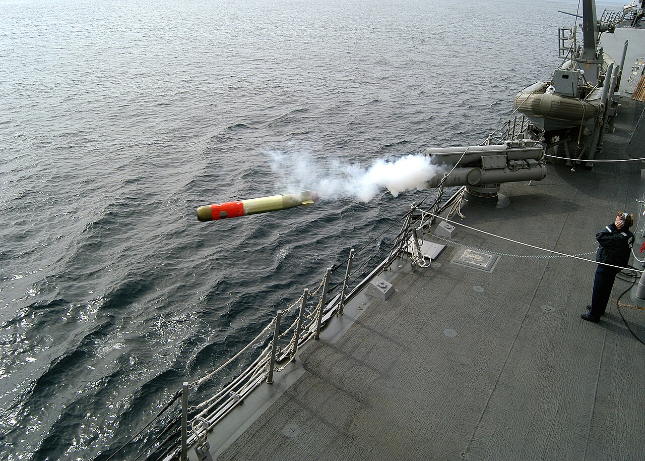A torpedo fired from a tube on the deck of a gray warship flies over the sea, trailing white smoke