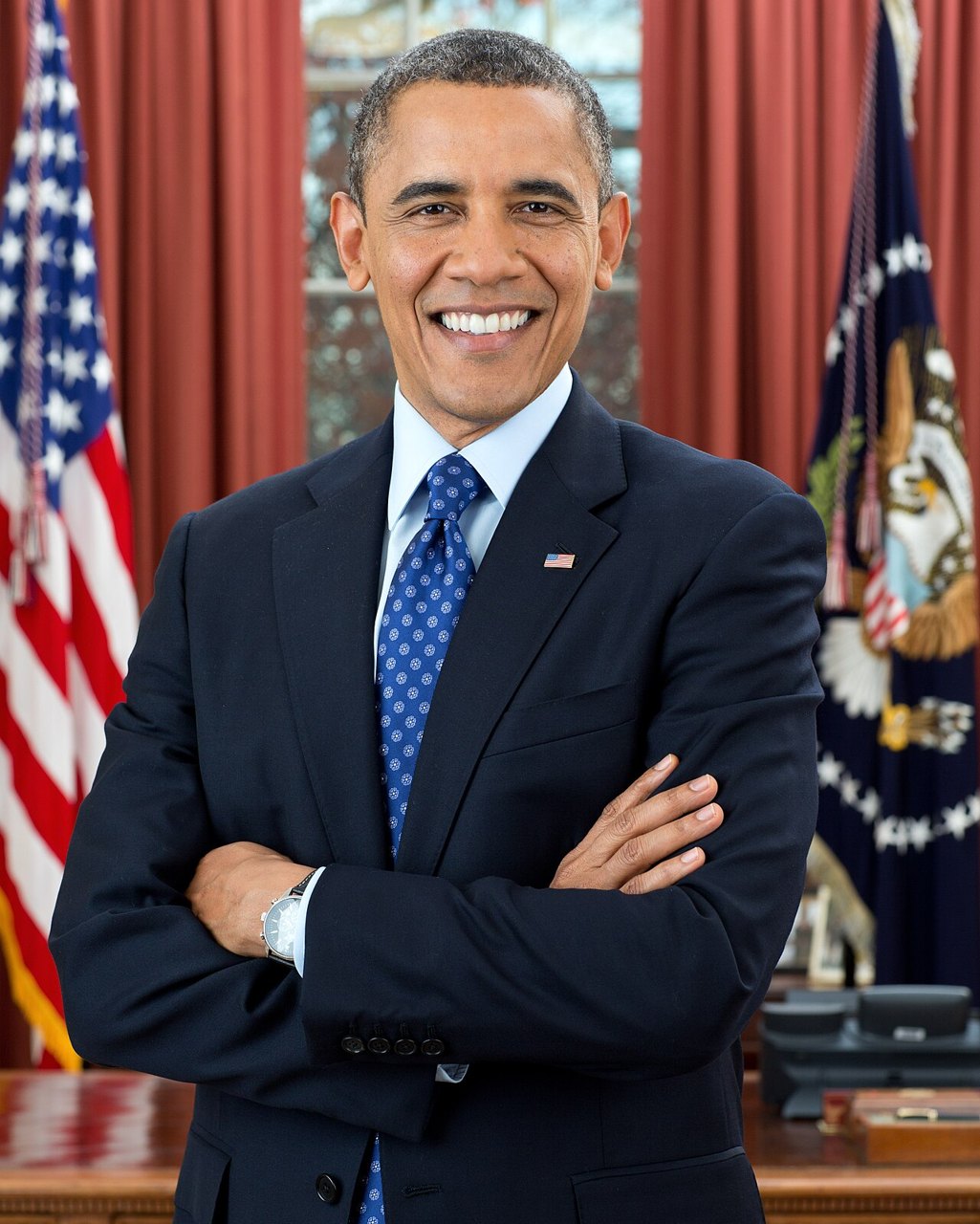 Barack Obama, smiling with his arms crossed, standing in the Oval Office