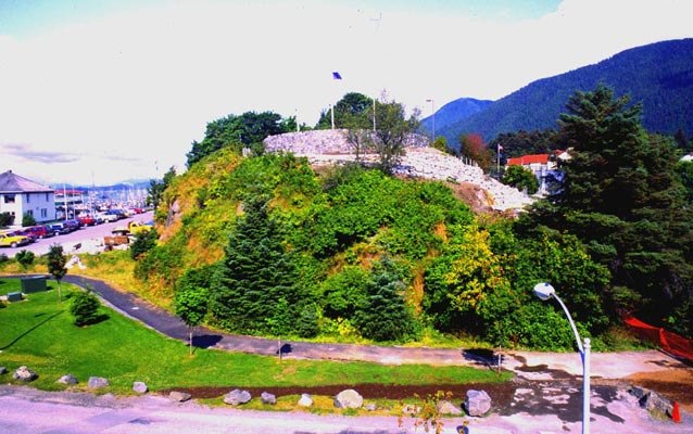 A green, tree-covered hill with a stone wall and flagpole on top, beside a road and a harbor in Sitka