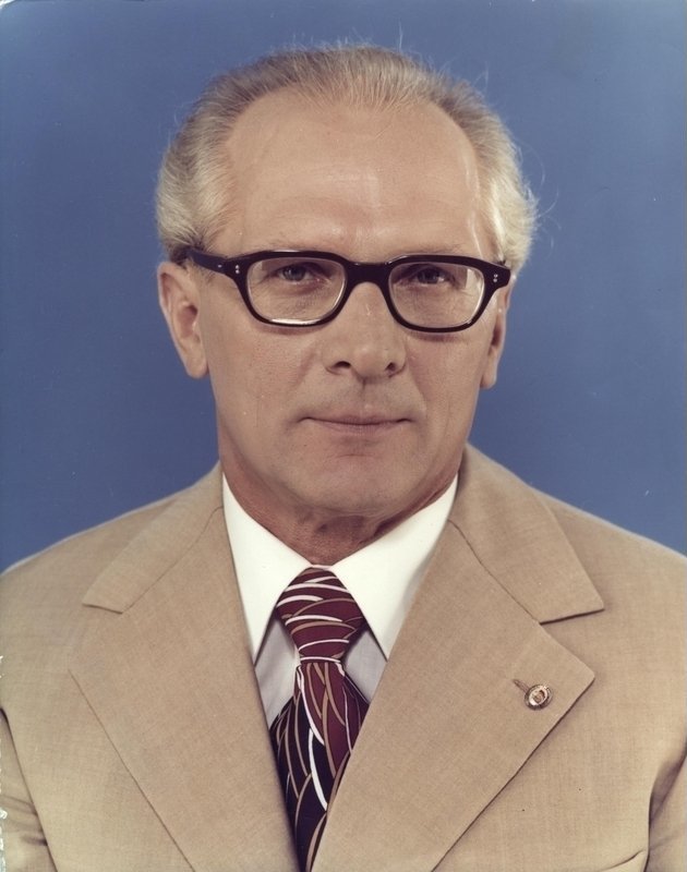 Studio portrait of a man with gray hair and dark-rimmed glasses wearing a tan jacket, white shirt and striped tie