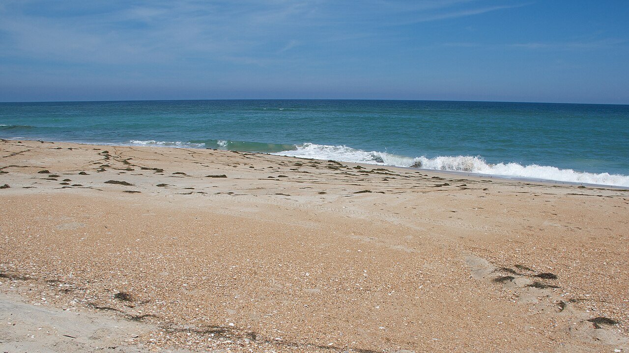Small waves break on an empty sandy beach under a blue sky with thin clouds