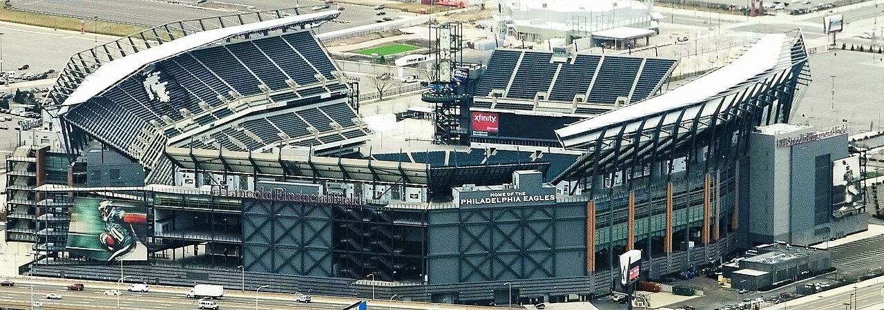 Aerial view of a large football stadium with dark blue seats and a sign reading Home of the Philadelphia Eagles, beside roads and parking lots