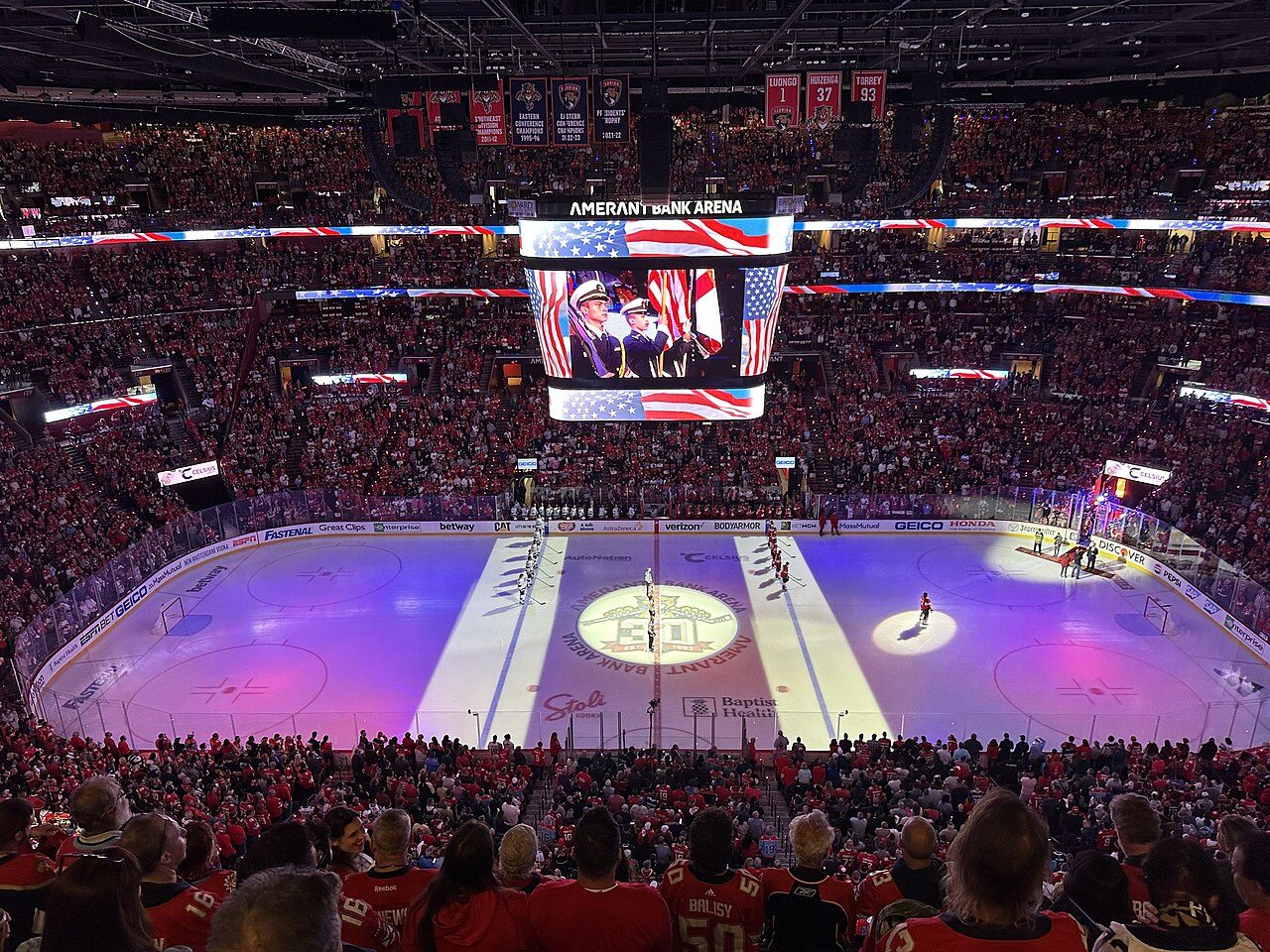 An arena packed with fans in red around a lit-up hockey rink, with a large video board hanging above the ice
