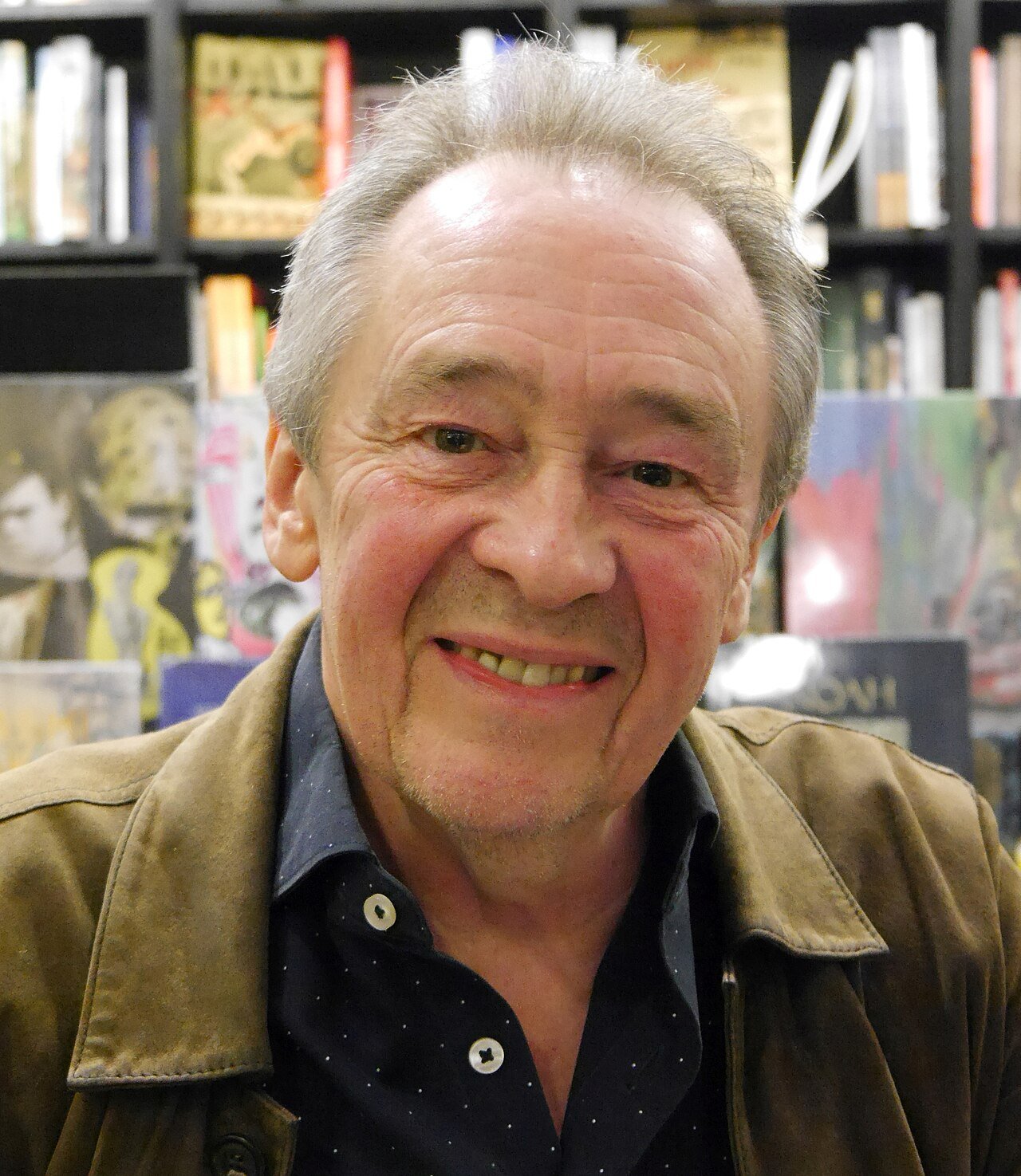 Paul Whitehouse, with grey hair and a tan jacket, smiles in front of shelves of books