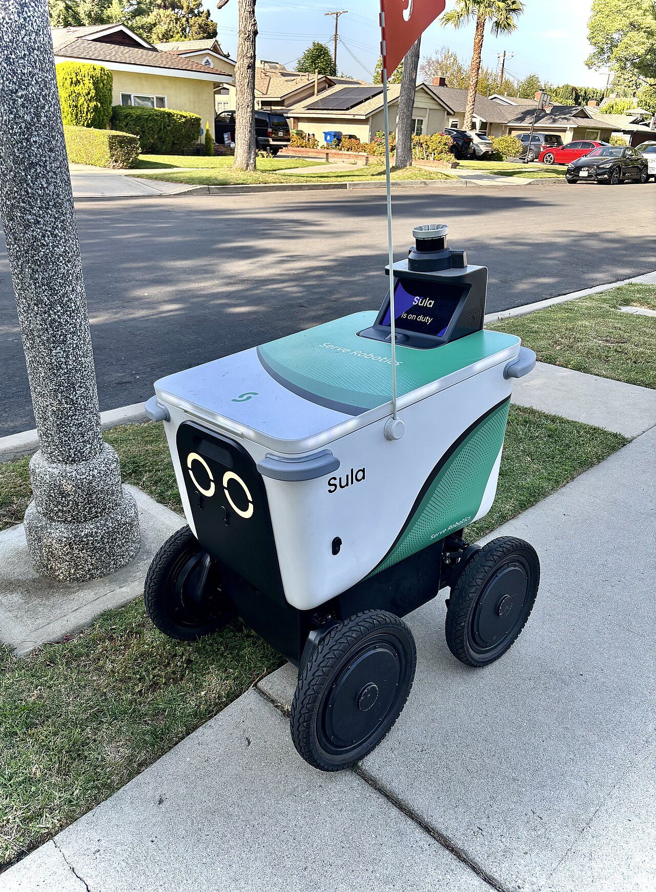 A small white and green four-wheeled delivery robot with a flag on top waits on a sidewalk beside a street, with palm trees in the background