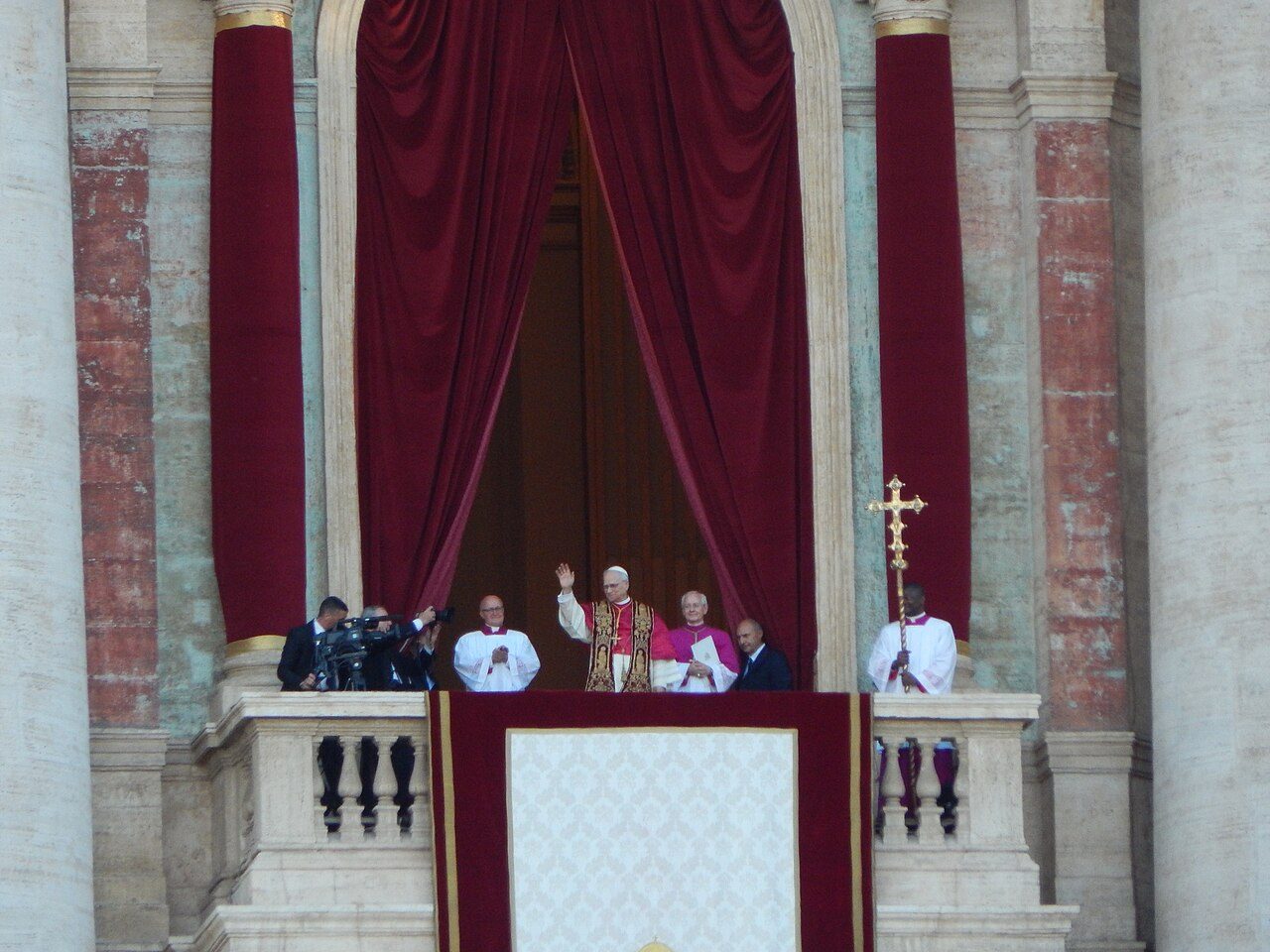 A pope in white and red vestments raises his hand to wave from a balcony draped with deep red curtains, flanked by clergy and a man holding a tall gold cross