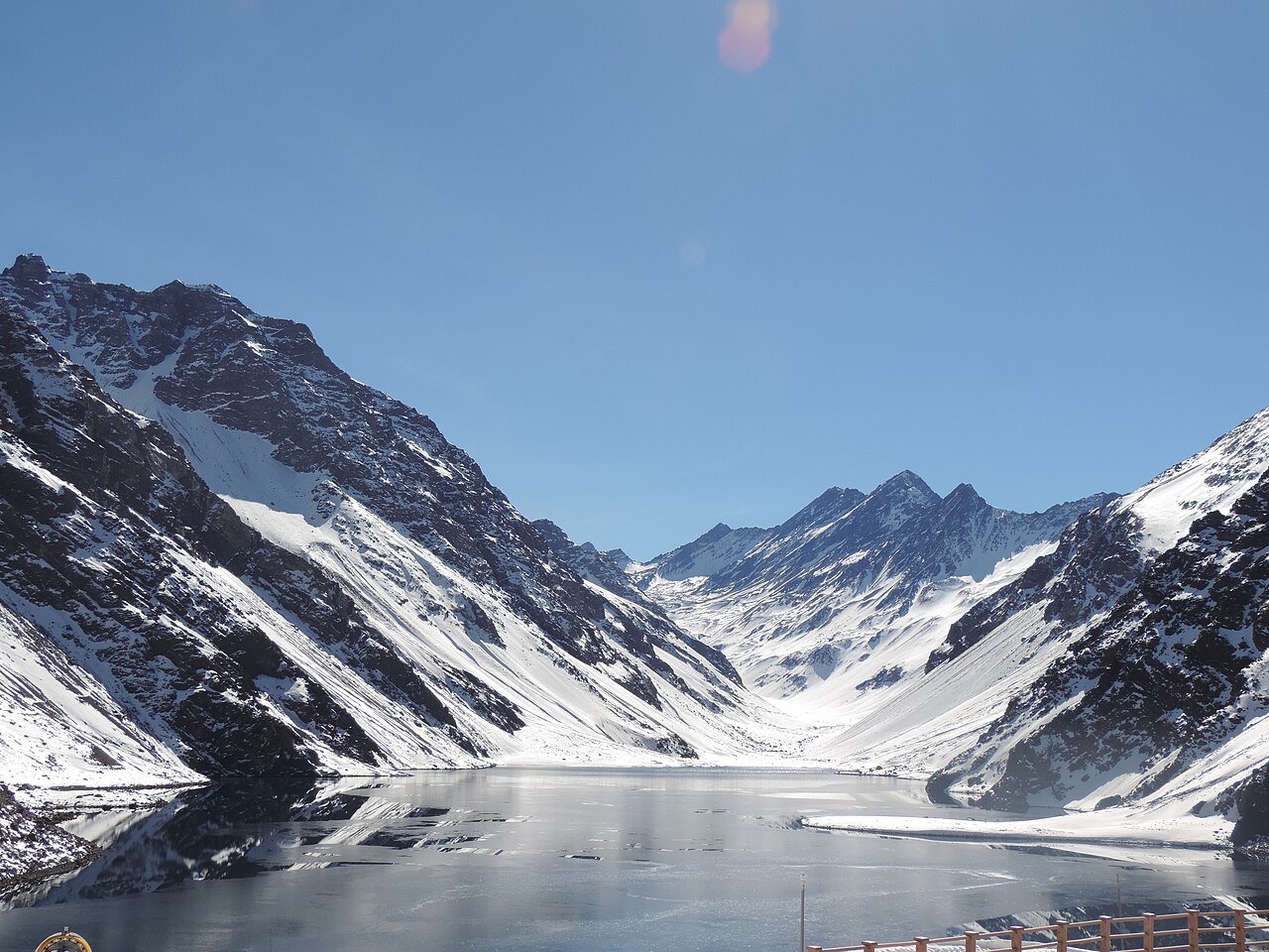 A partly frozen mountain lake sits below steep snow-covered peaks under a clear blue sky