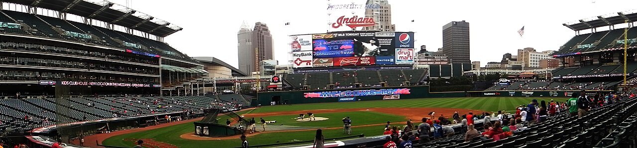 A panoramic view of a baseball stadium with a green field, a large scoreboard topped by old "Indians" signage, and city towers behind the outfield during batting practice