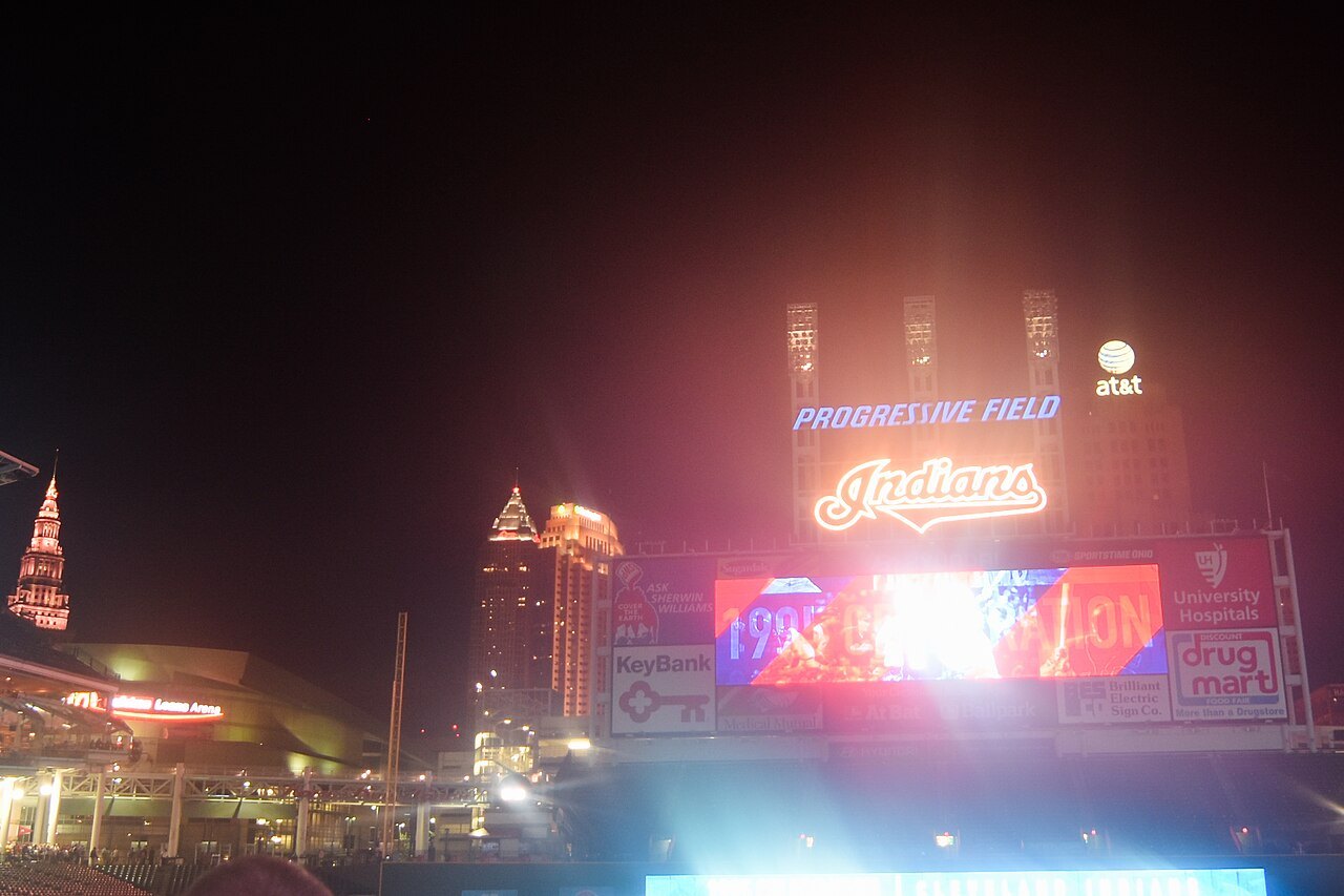 The lit scoreboard at Progressive Field at night, with downtown Cleveland towers behind it