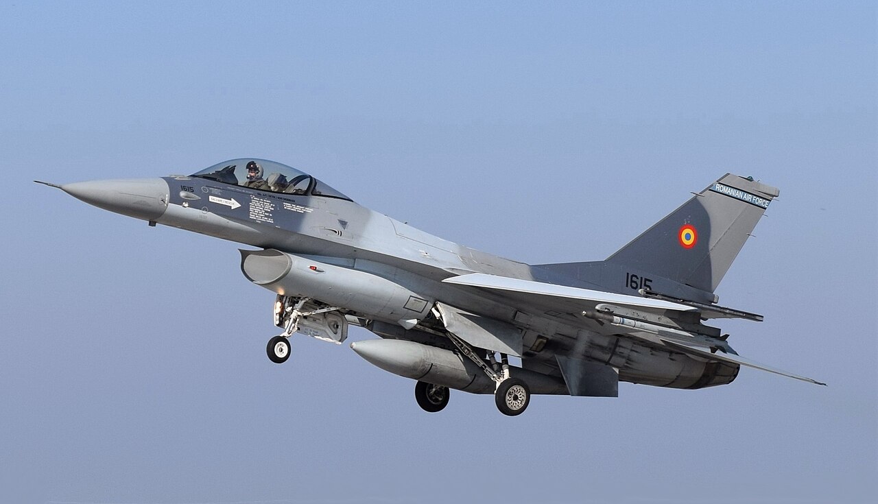 A gray fighter jet with its landing gear down flies low against a pale sky
