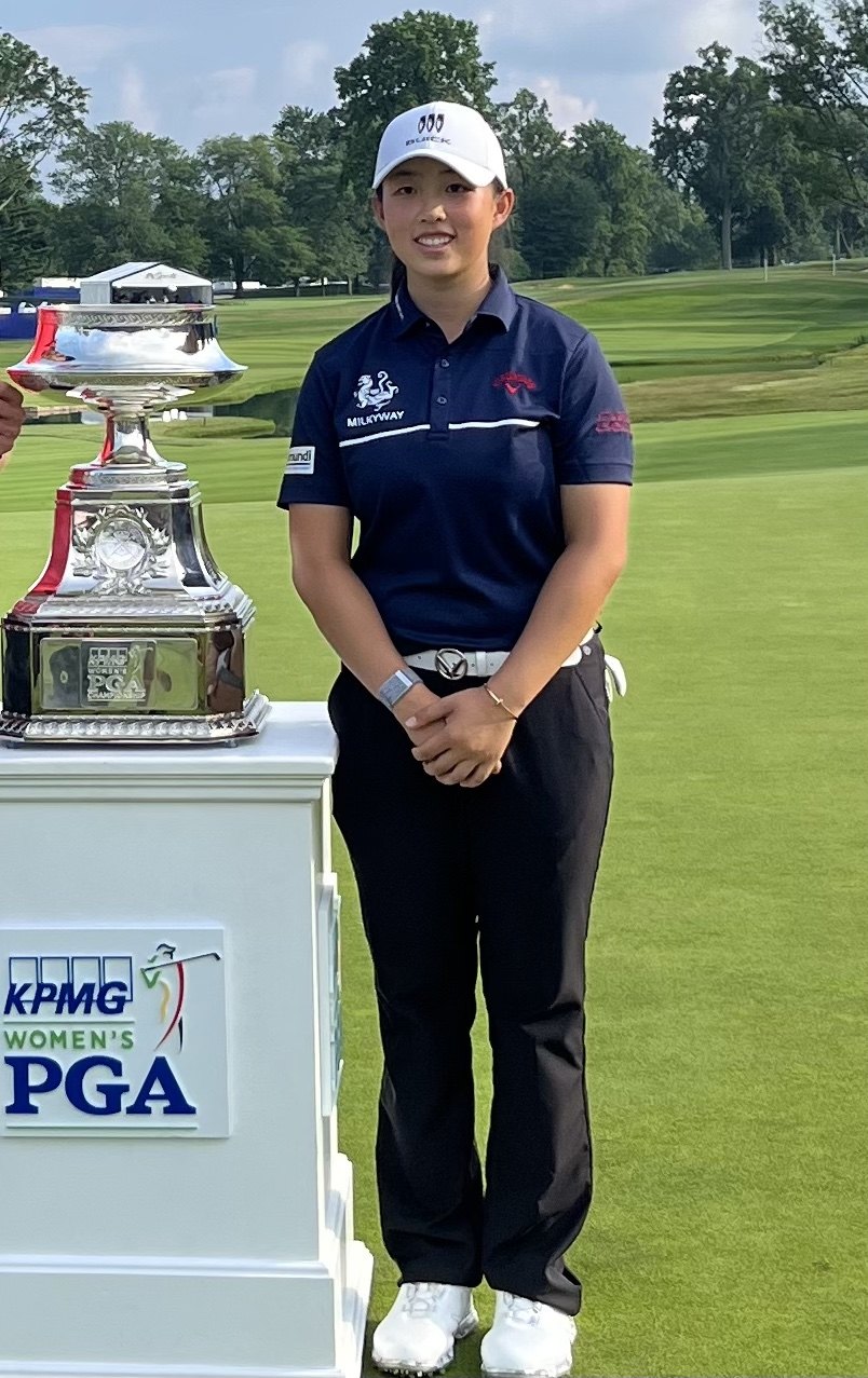 Golfer Ruoning Yin in a white cap and navy shirt standing beside a large silver trophy on a golf course