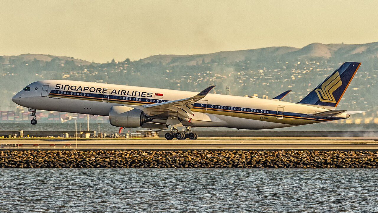 A white Singapore Airlines jet with a dark blue and gold tail lifts off from a runway beside the water, with hills in the background