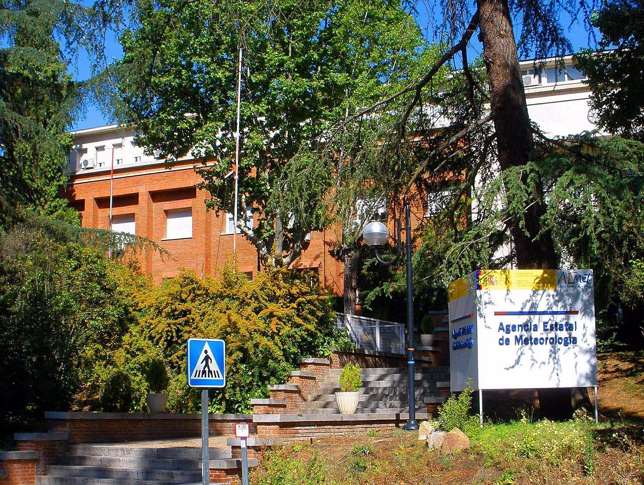 A red-brick building behind trees and shrubs, with a sign reading Agencia Estatal de Meteorología beside a set of steps
