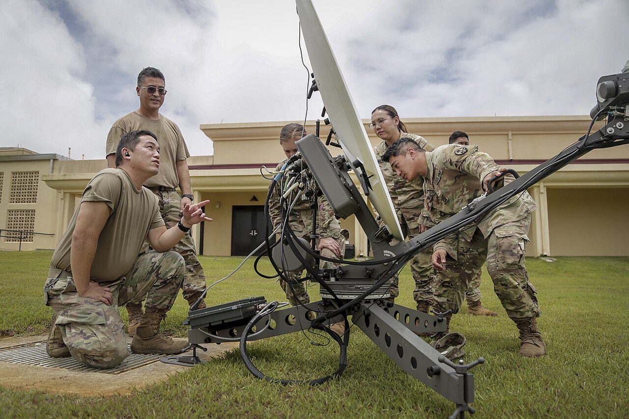 Service members in camouflage uniforms gather around a portable satellite dish on a tripod on a grassy lawn, with a low tan building behind them under a cloudy sky