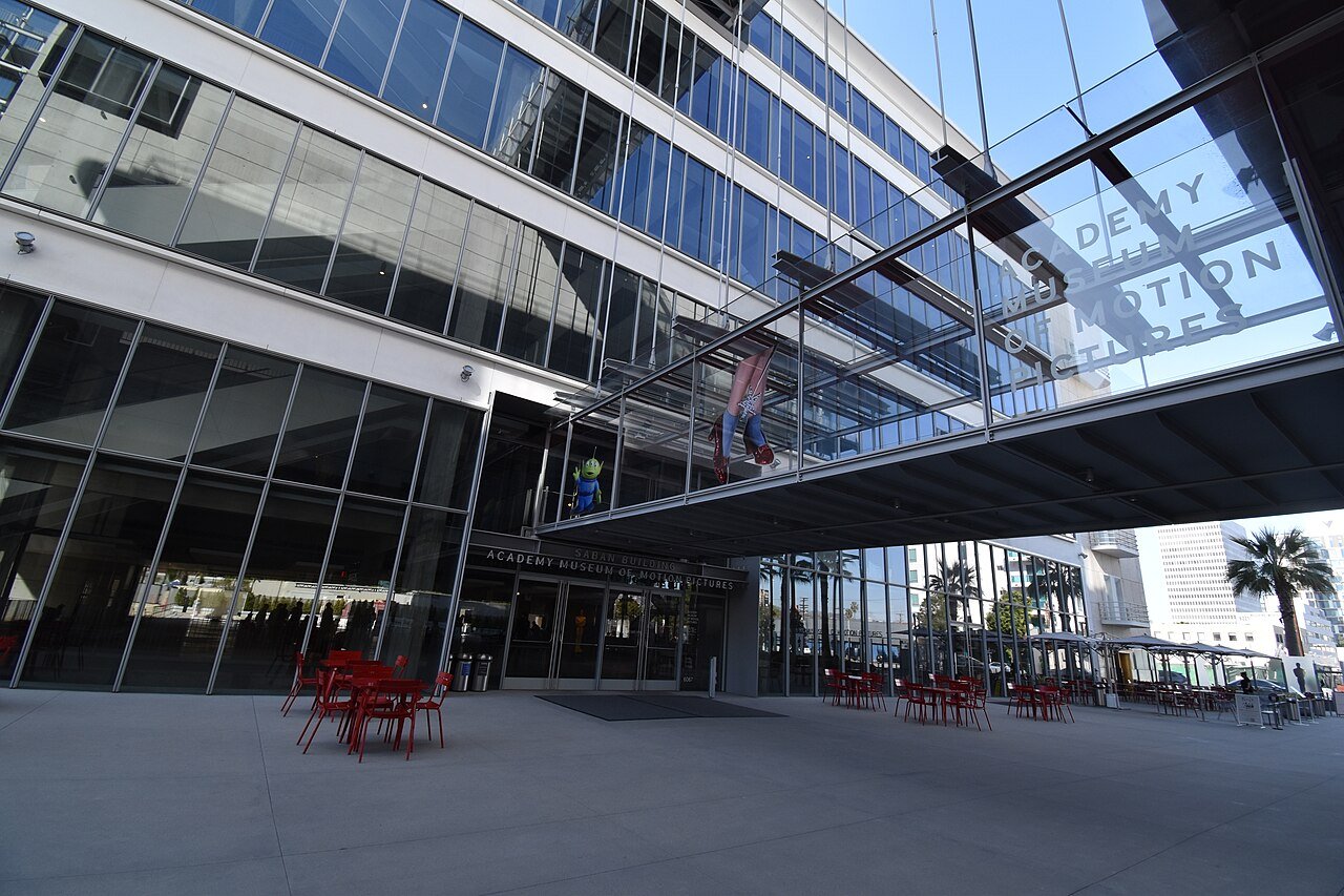 The glass entrance of the Academy Museum of Motion Pictures, with its name over the doors and on a glass bridge overhead, where a hanging Spider-Man figure and a small green alien toy figure are on display above red chairs and tables on a plaza