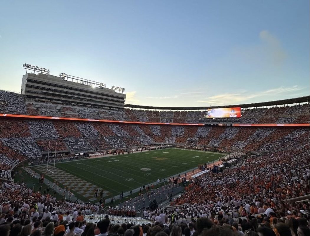 A huge football stadium packed with fans wearing orange and white in a checkered pattern, with the green field and evening sky above