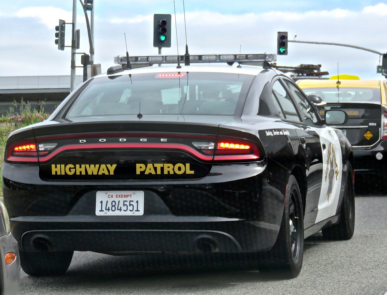 The rear of a black and white Dodge Charger patrol car marked Highway Patrol, stopped at an intersection with traffic lights overhead