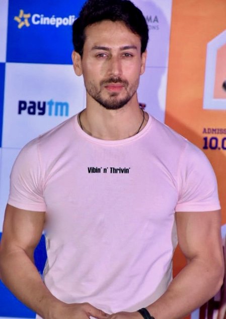 A man with short dark hair and a beard, wearing a pink T-shirt, stands in front of a sponsor backdrop at a promotional event
