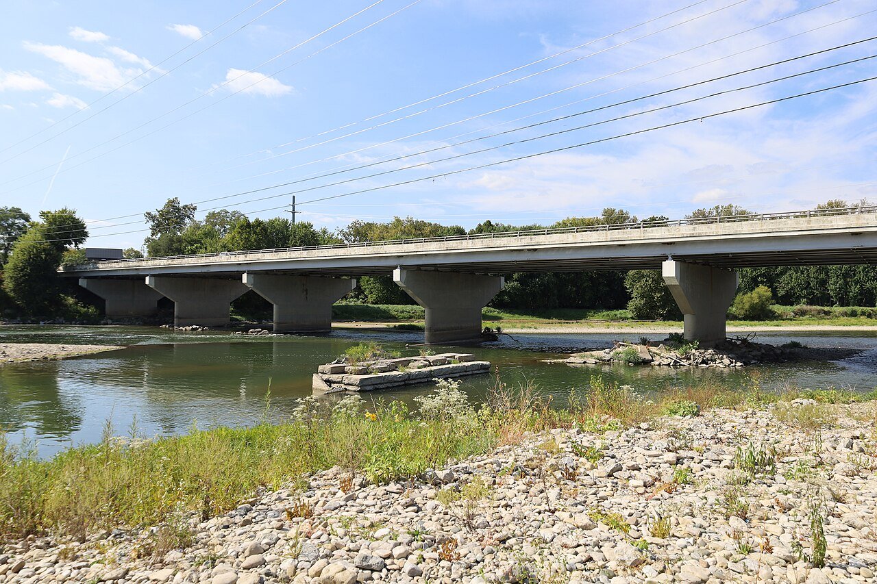A concrete road bridge crosses a shallow river with a rocky bank under a blue sky