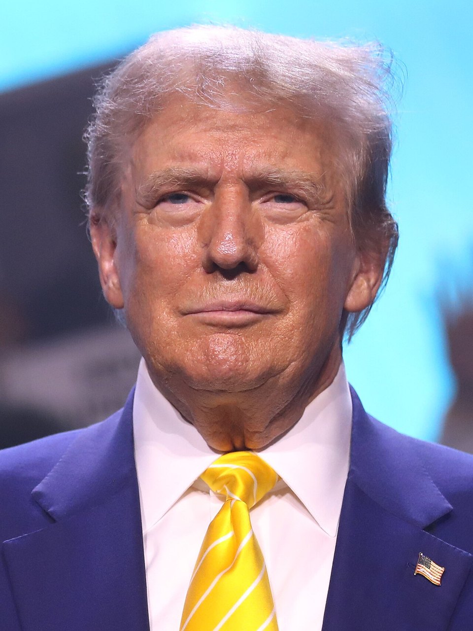 Donald Trump in a dark suit and yellow tie, against a blue background
