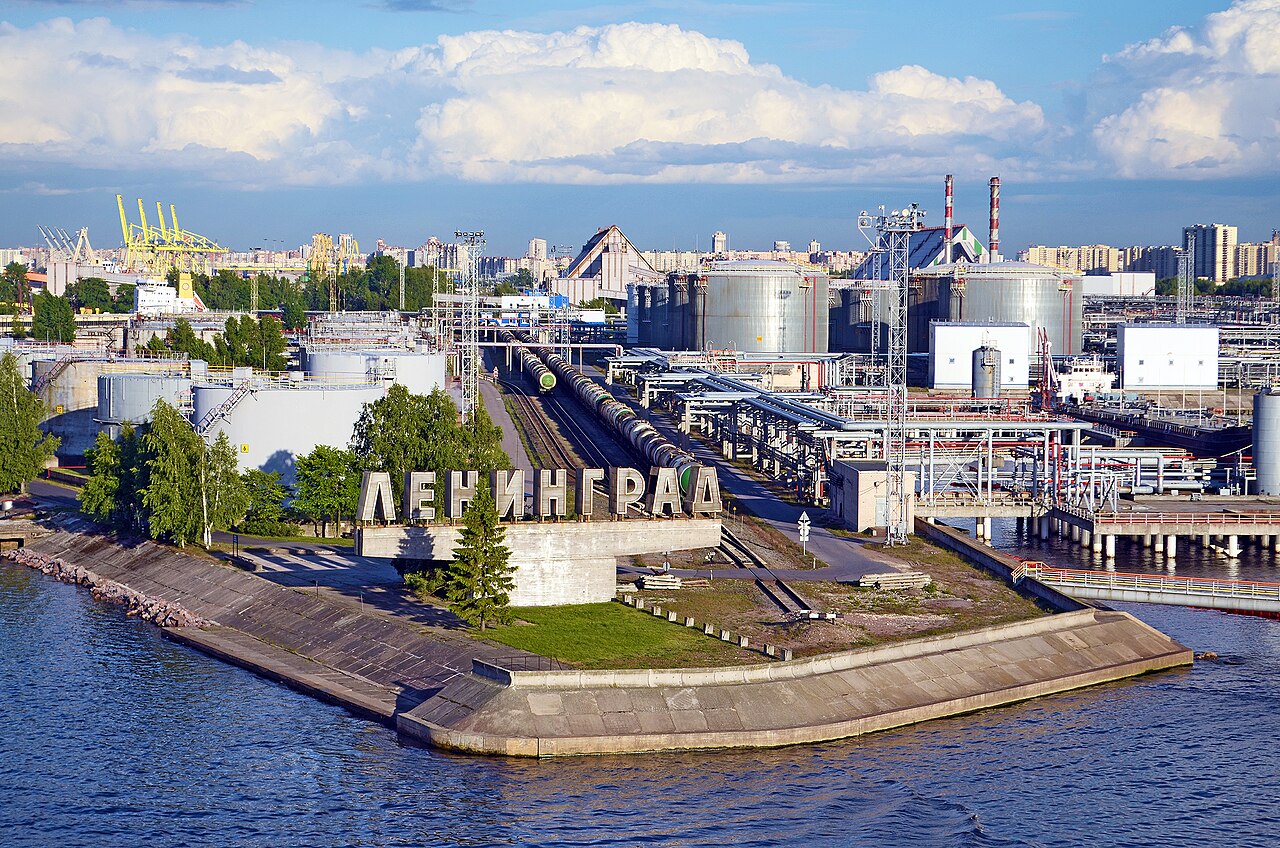 Oil storage tanks, rail tank cars and cranes at a busy port beside the water, with a large sign spelling Leningrad in Russian