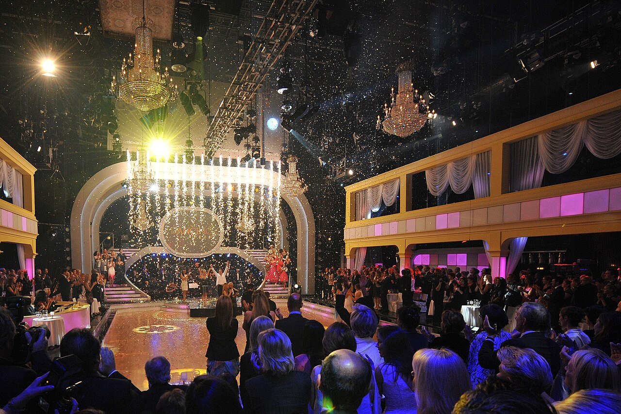 A glittering TV ballroom with chandeliers and falling confetti over a dance floor as an audience watches