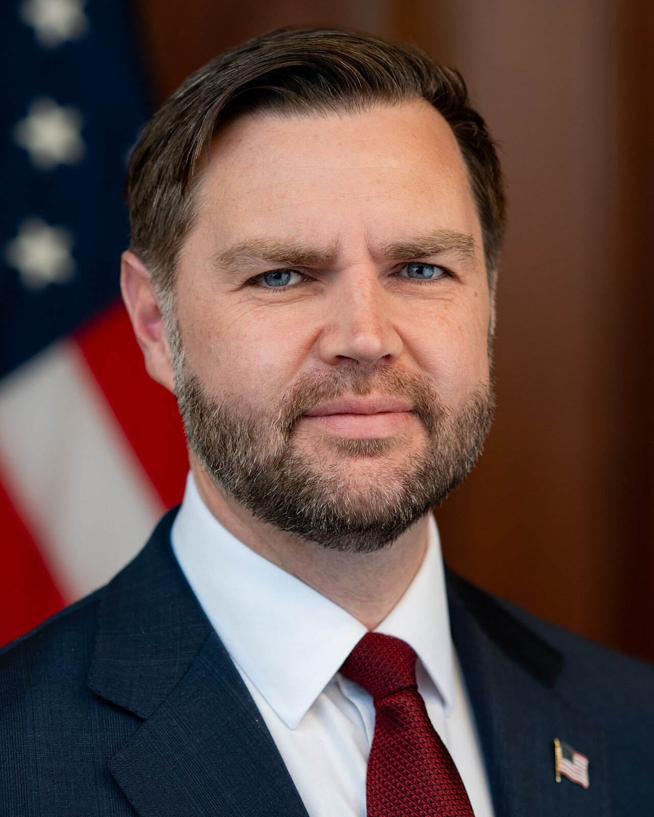 Vice President JD Vance in a dark suit and red tie stands in front of an American flag in an official portrait