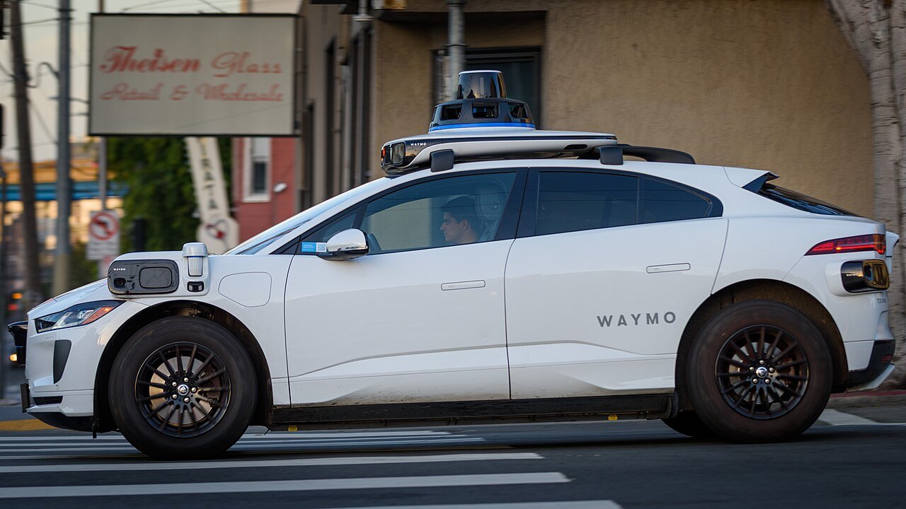 A white Jaguar SUV with sensors mounted on its roof drives along a city street past storefronts