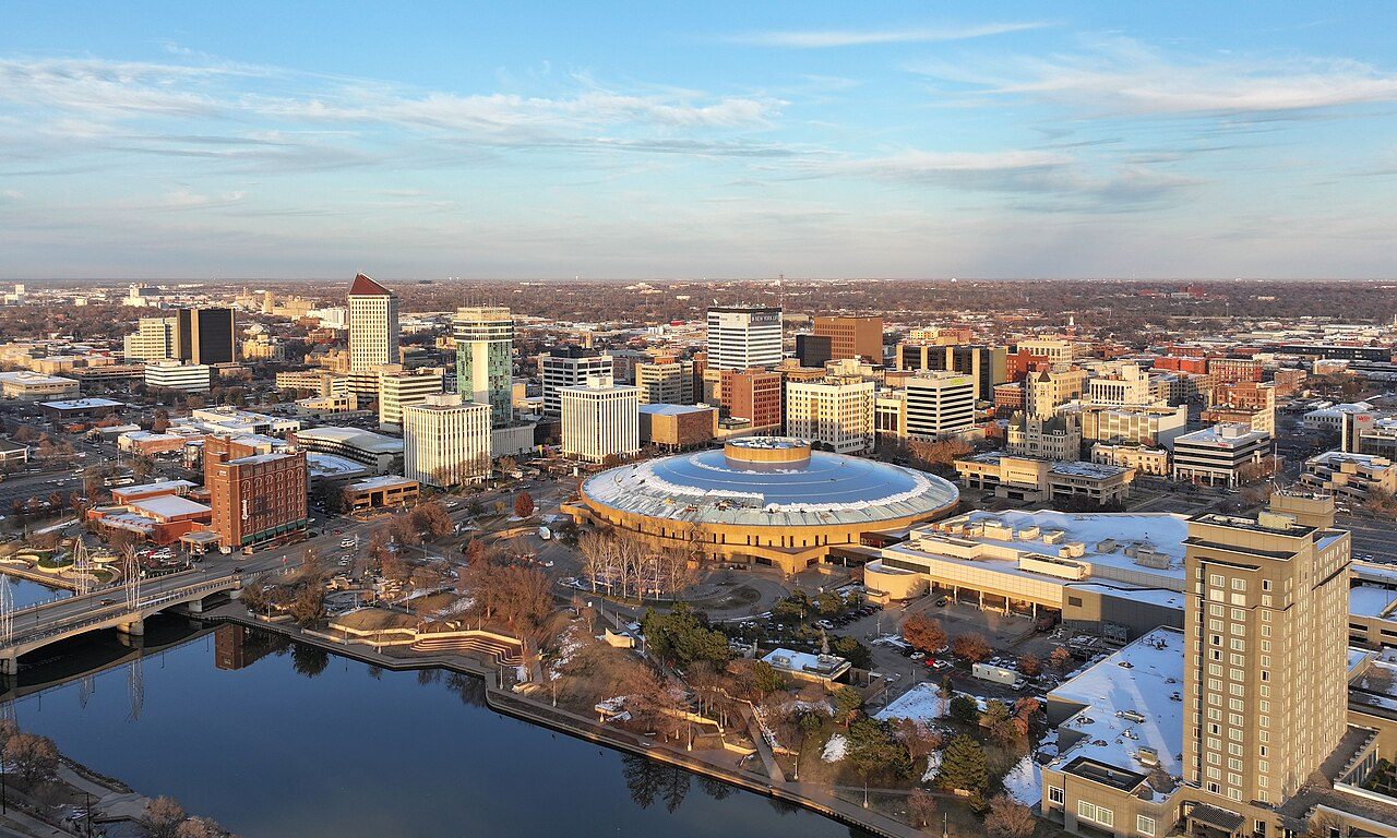 An aerial view of downtown Wichita at sunset shows a round domed arena beside a river, with office towers, hotels and low buildings spreading out under a pale blue sky