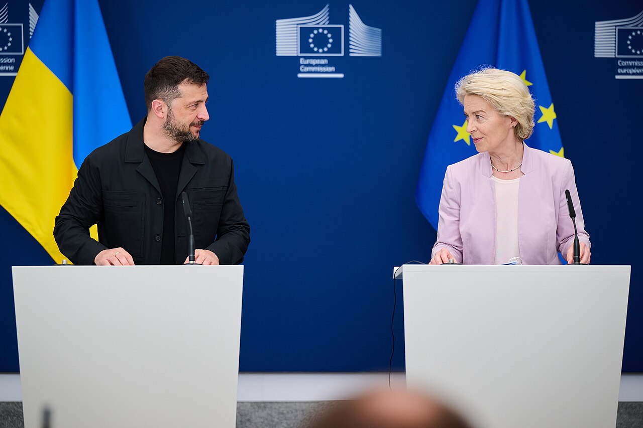 Two leaders stand at separate white podiums in front of blue flags, a man in a dark sweater on the left and a woman in a pink jacket on the right