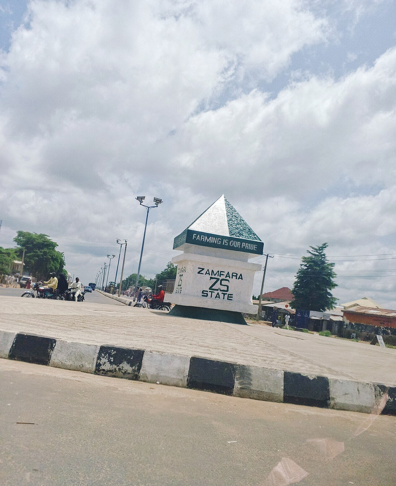 A white pyramid-topped monument with a Zamfara State sign stands at a roundabout on a paved road under a cloudy sky