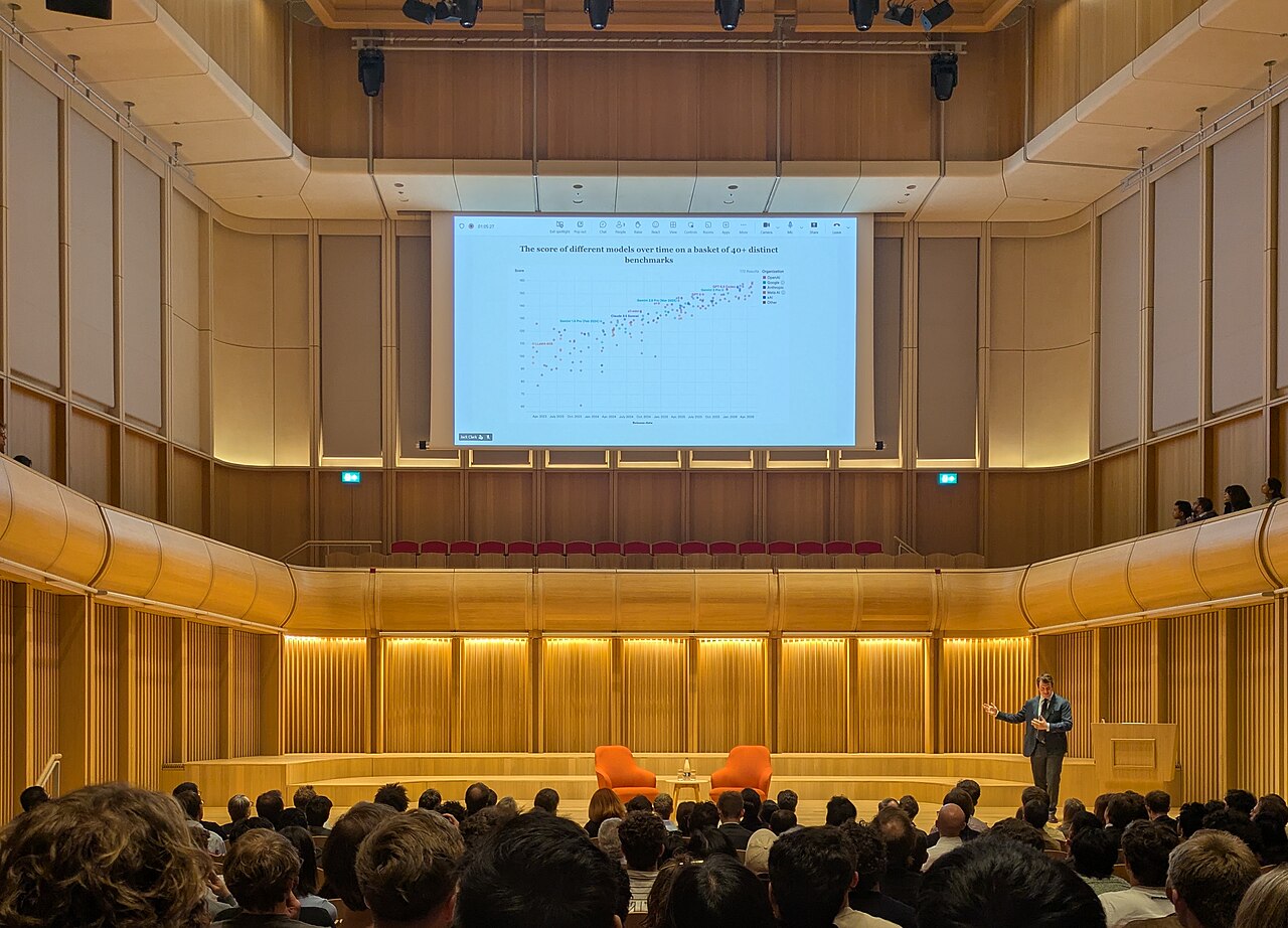 A man speaks on a stage in a large wood-paneled concert hall in front of a seated audience and a big screen showing a chart